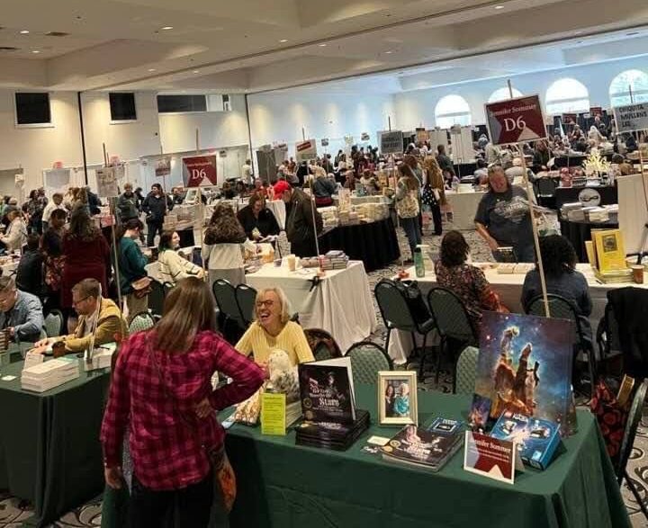 A large convention room with tables filled with books. Authors are greeting readers.
