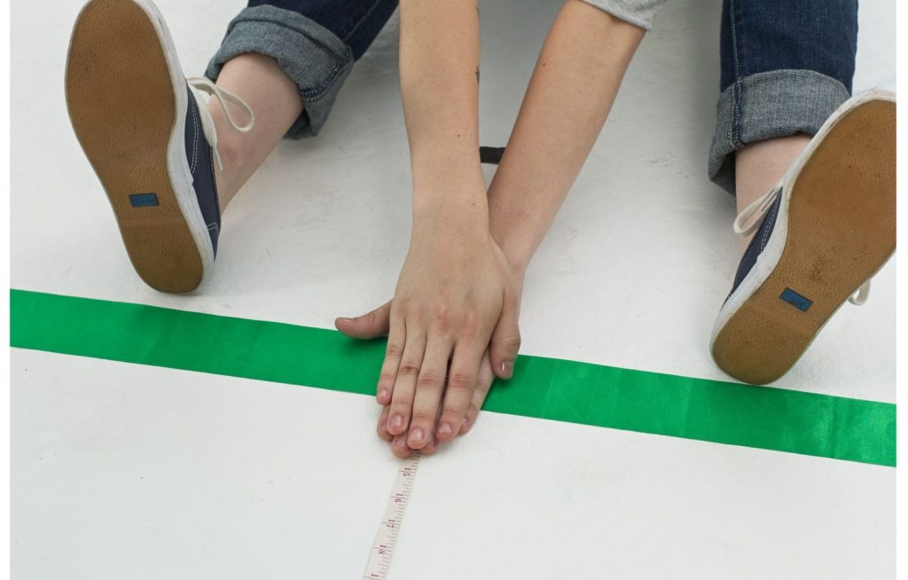 A green ribbon with fingertips and toes reaching past. A tape measure is on the floor to measure how far the fingertips and reach.